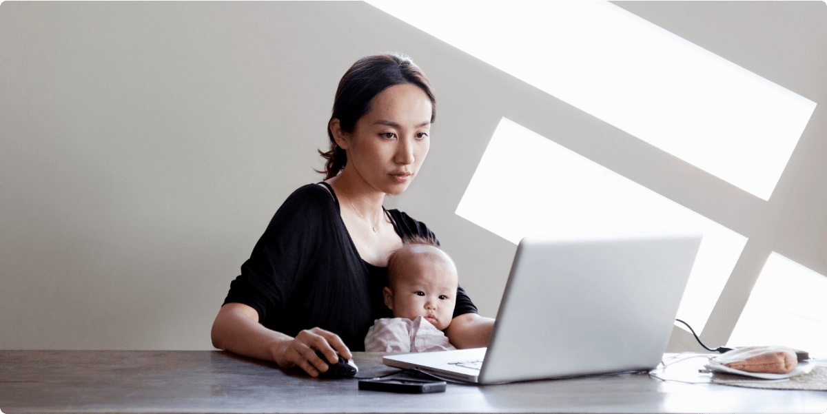 Una madre con su niño pequeño en su regazo, trabajando en un portátil. La luz del sol entra a raudales detrás de ella.