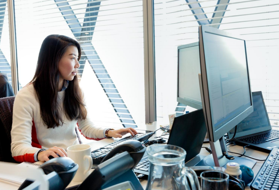 Donna seduta alla scrivania di un ufficio davanti a un computer portatile e a un monitor.